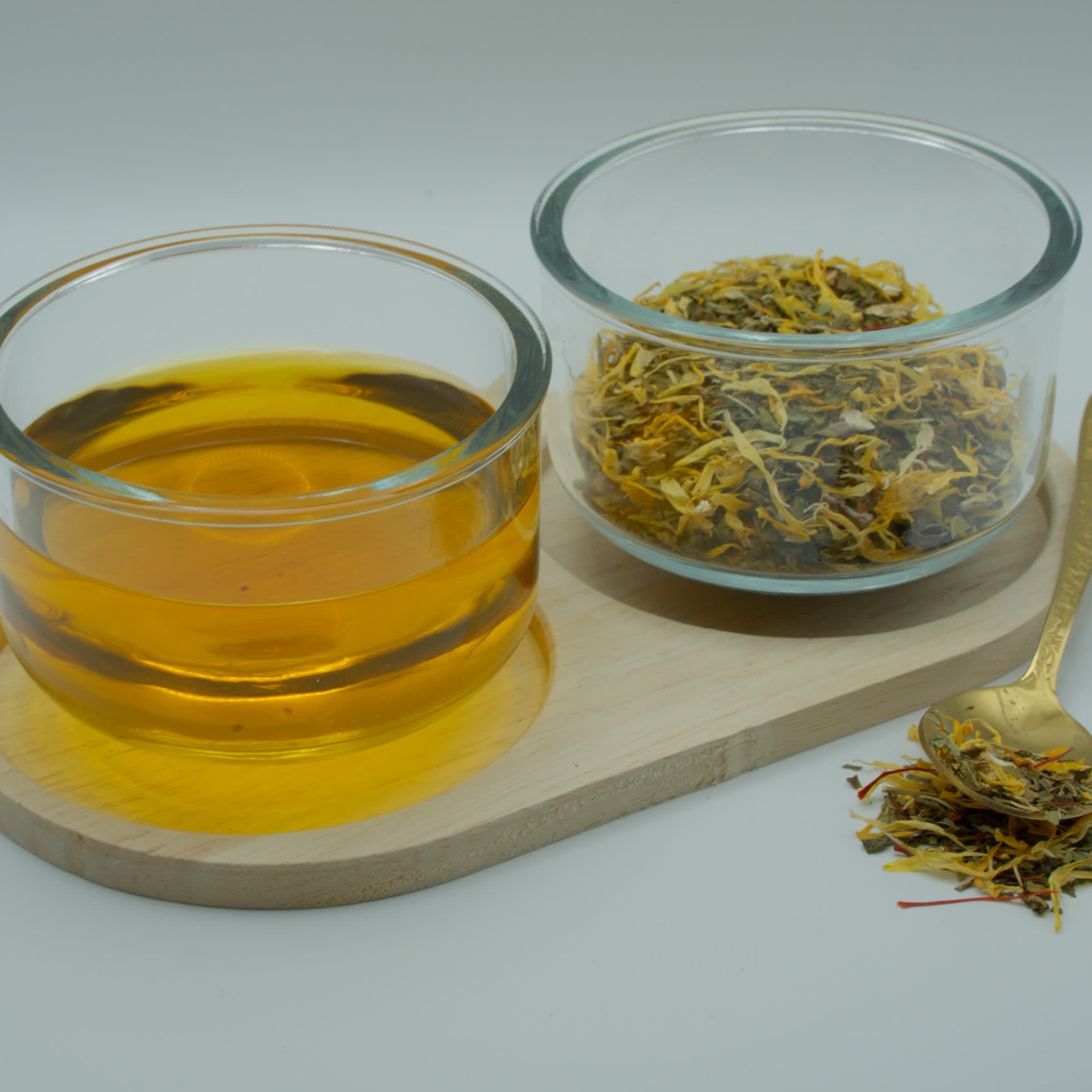 A glass bowl of brewed amber tea beside a bowl of dried mint, cacao nibs and yellow calendula petals on a wooden board