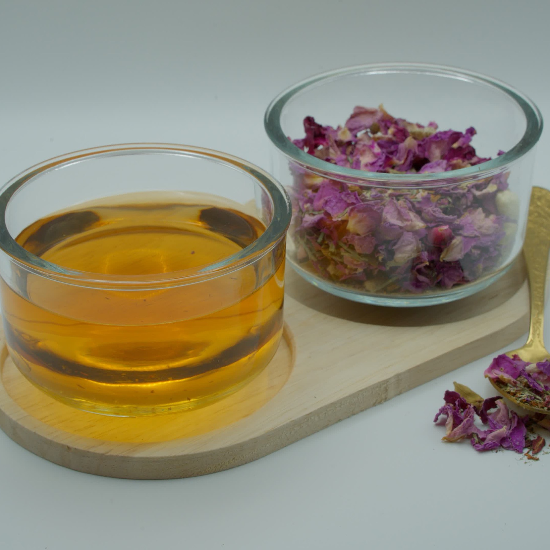 A glass bowl of brewed amber tea beside a bowl of dried pink rose petals on a wooden board, with a gold spoon